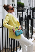 Lalage Beaumont female in a yellow blazer and white pants holding ninette orinoco Handbag against a black iron fence.