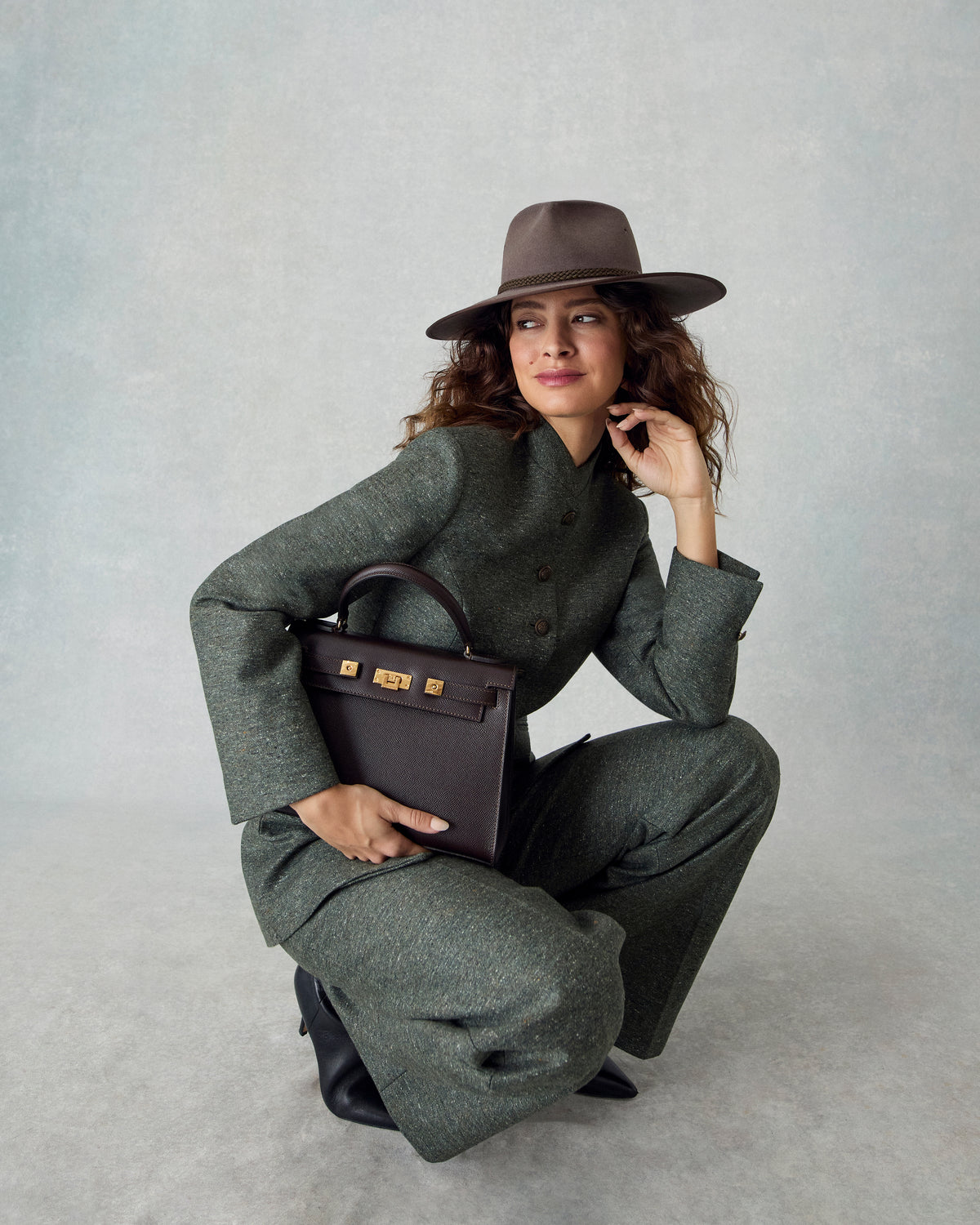 A woman crouches in a studio wearing an olive green tailored suit, a brown wide-brim hat, and holding a dark brown structured handbag with gold hardware against a light gray background.