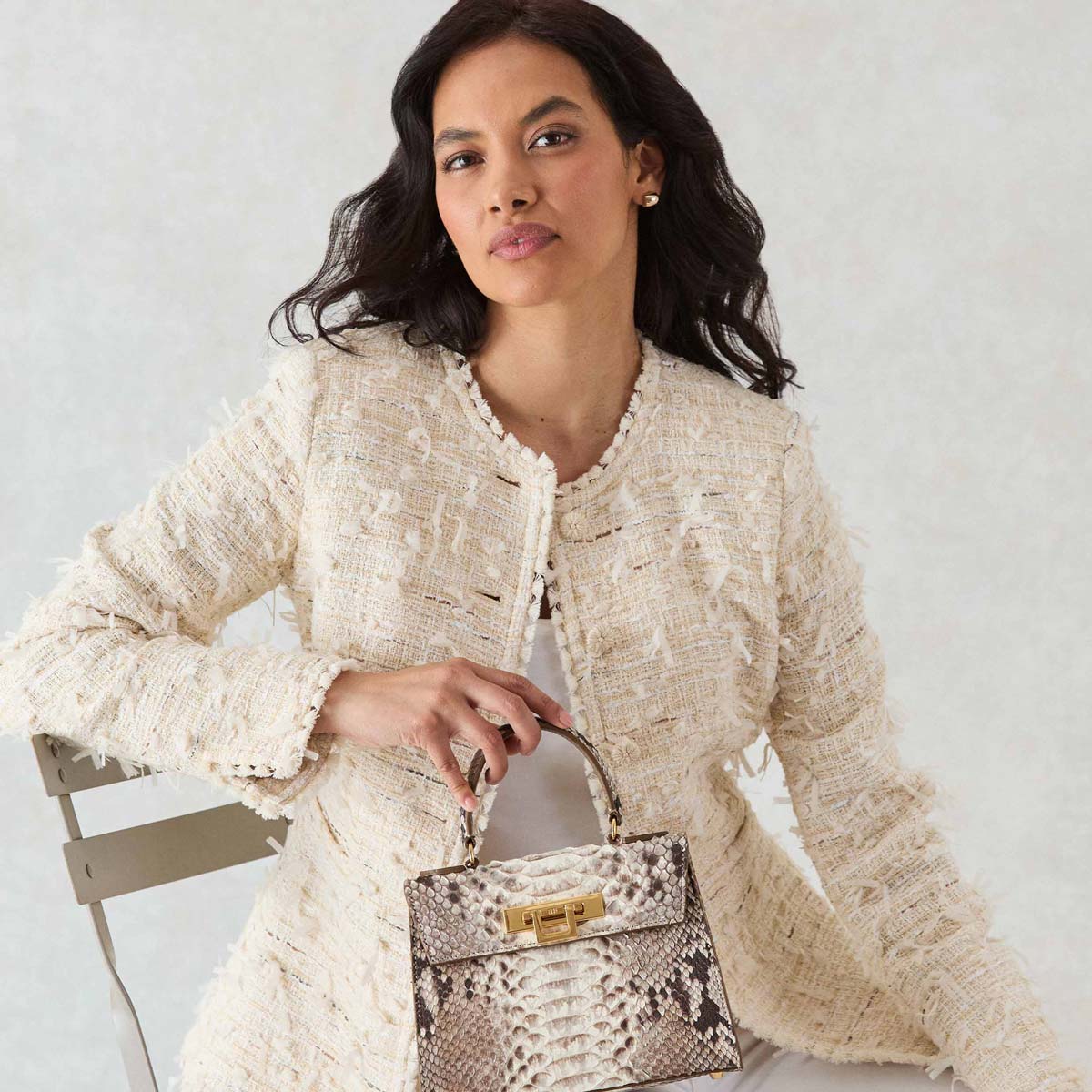 Woman holding a Lalage Beaumont Maya Midi Pythonskin Handbag wearing a textured beige jacket against a light background


