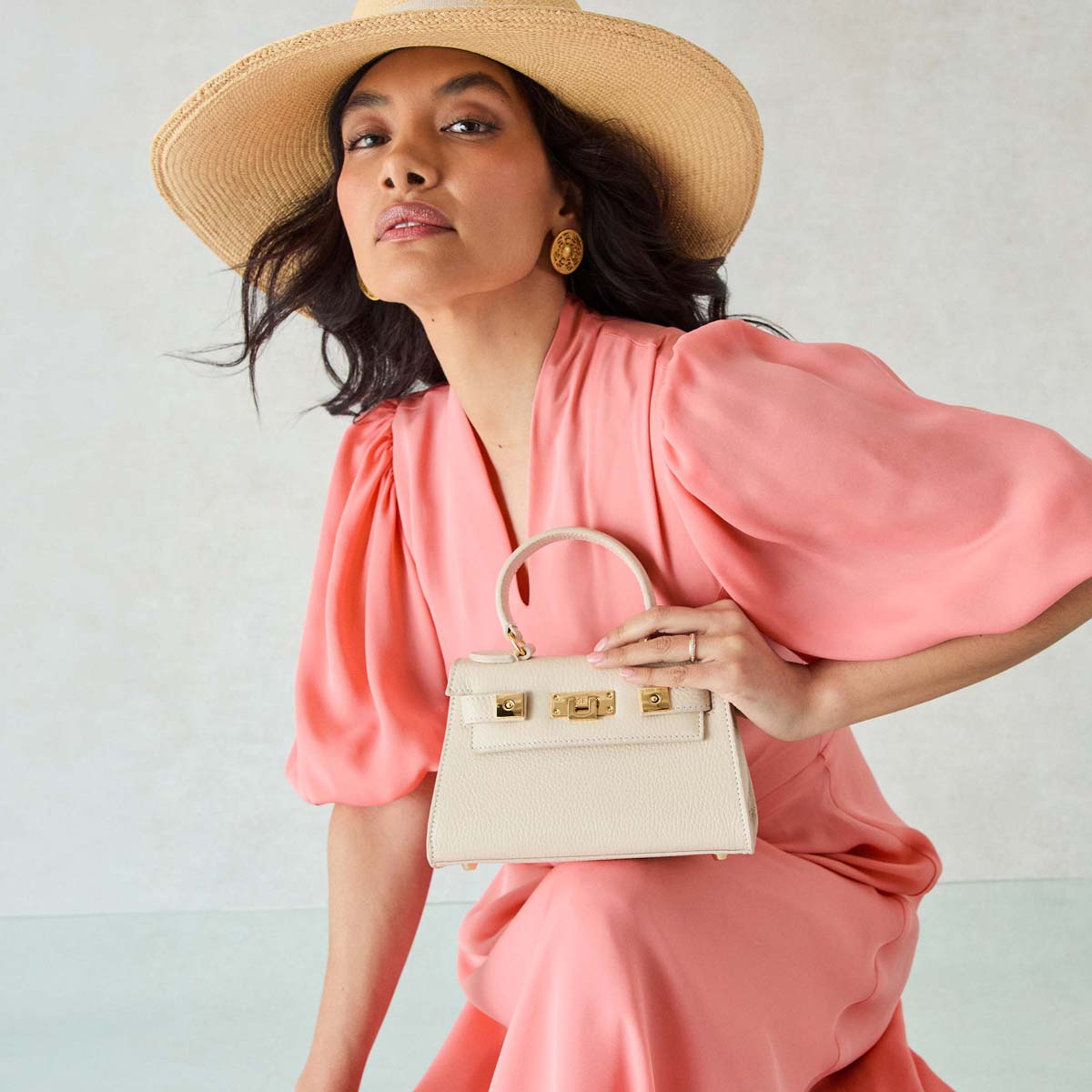 Woman in a pink dress and straw hat holding a Lalage Beaumont cream handbag against a light background.