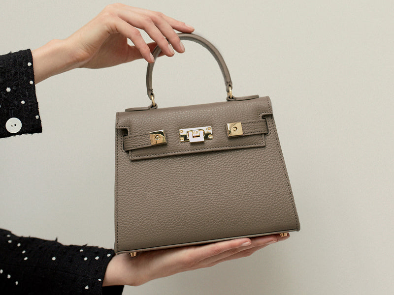 A taupe colored, medium-sized handbag made from grainy print Italian calf leather, featuring gold-plated hardware and a top handle with a detachable shoulder strap.