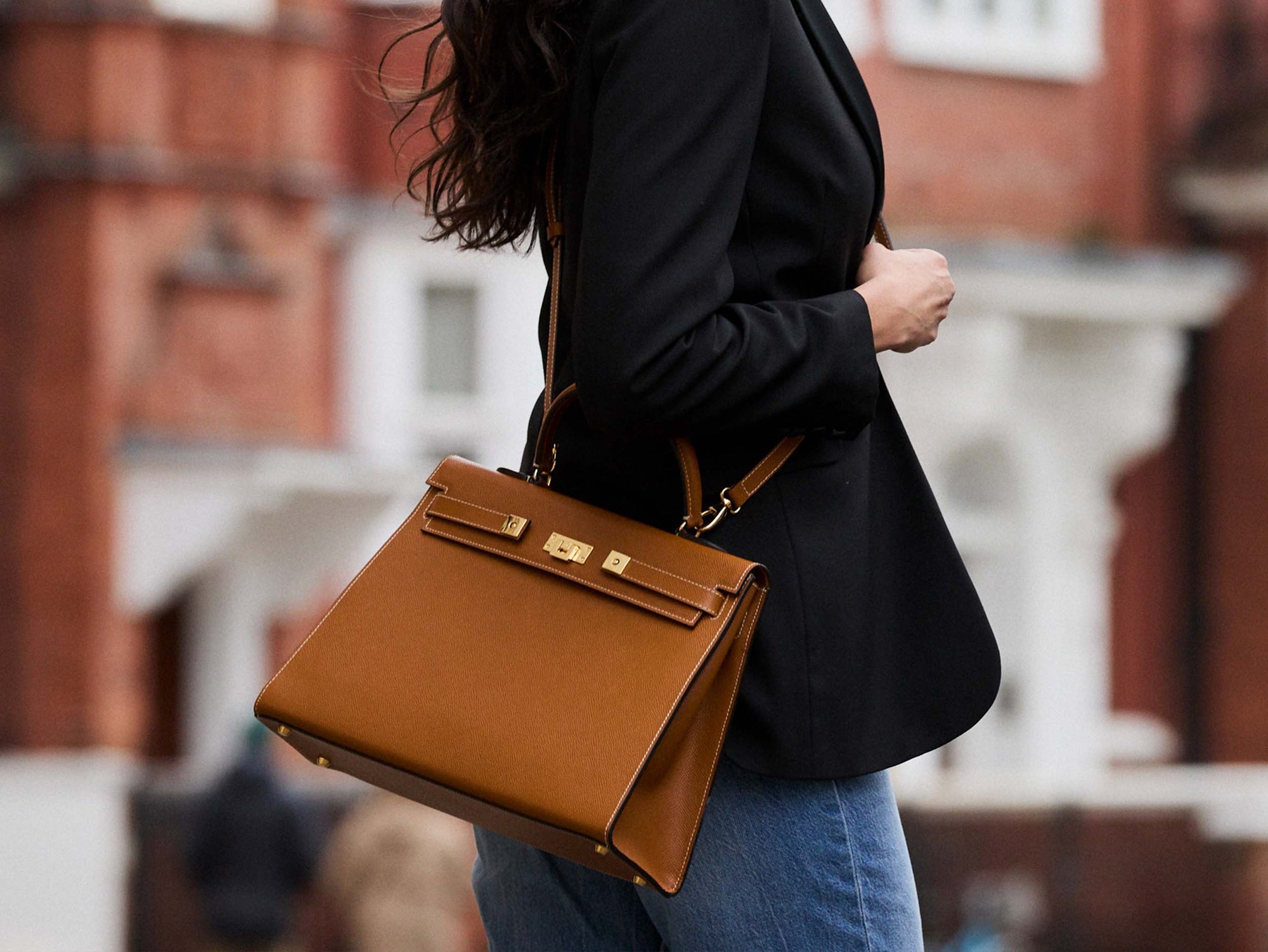 A large tan colored Italian calf leather handbag with pebble print texture, gold plated hardware, and a top handle with a detachable strap carried by a woman.