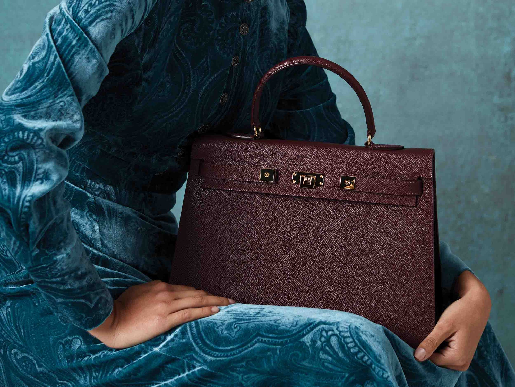 Person holding a large wine colored Italian calf leather handbag with pebble print texture, gold plated hardware, and a top handle with a detachable strap against a textured blue background