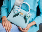 Women holding a medium-sized luxury Italian leather handbag in a soft aqua pale turquoise color with a grainy texture, featuring a top handle, a detachable shoulder strap and gold-plated hardware