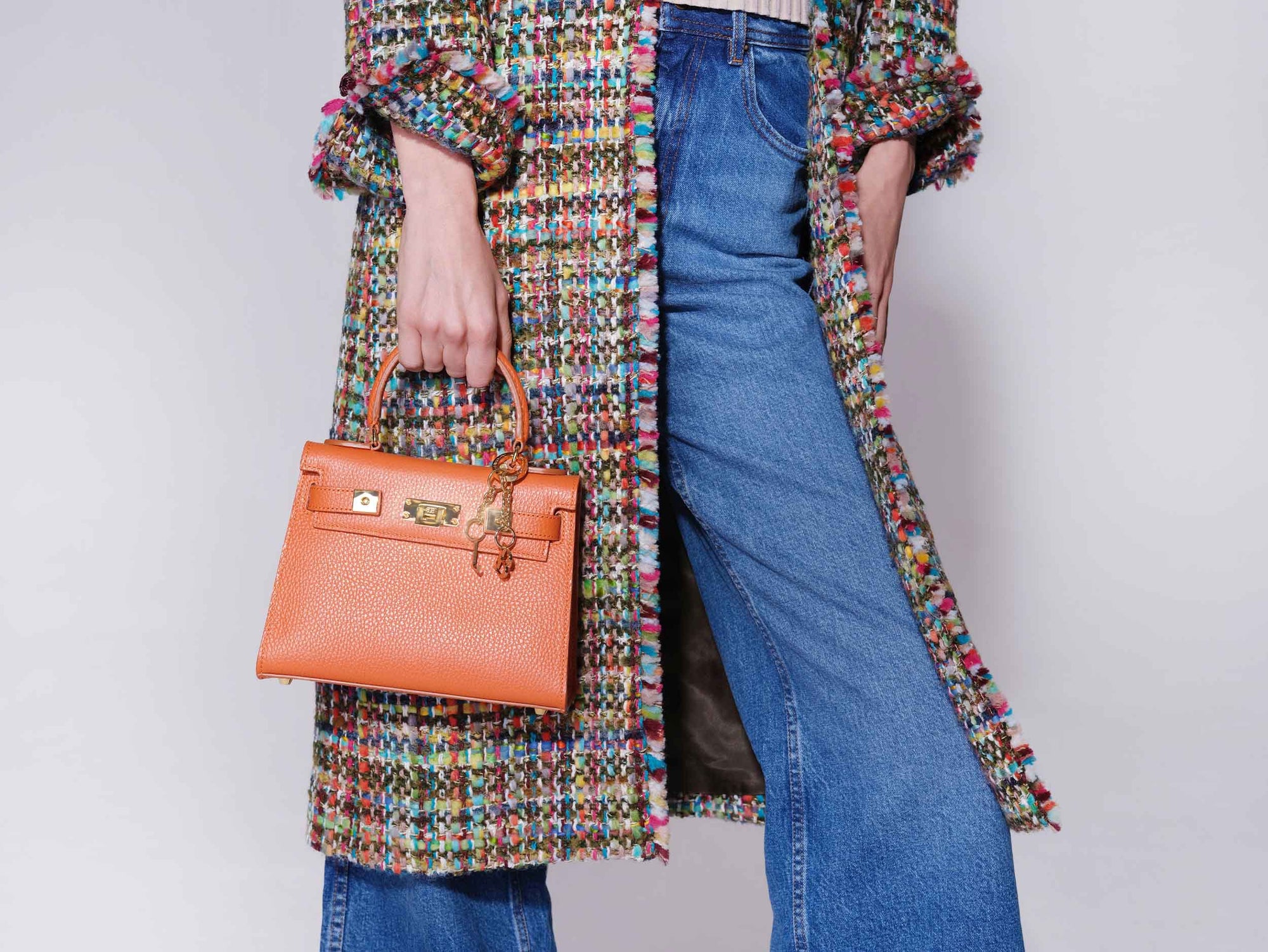Person wearing a colorful coat and blue jeans, holding an orange luxury handbag with a top handle and gold-plated hardware, made of Italian calf leather against a plain background