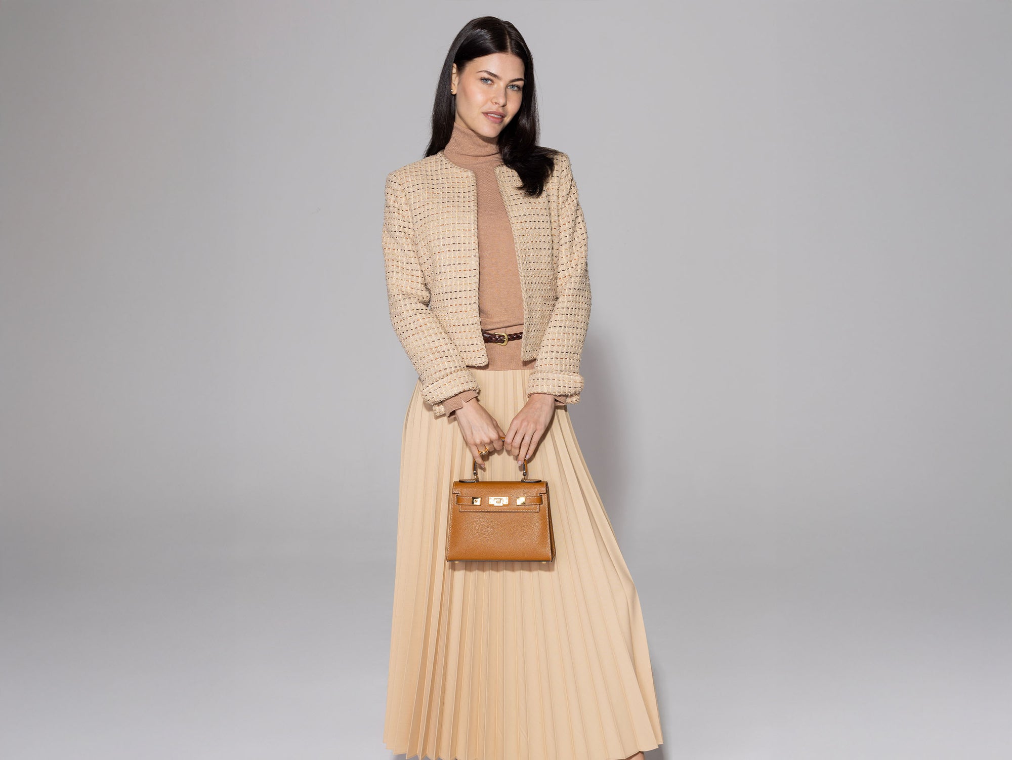 Woman holding a tan-colored, pebble print Italian calf leather handbag with gold-plated hardware, a top handle and a detachable strap against a gray background