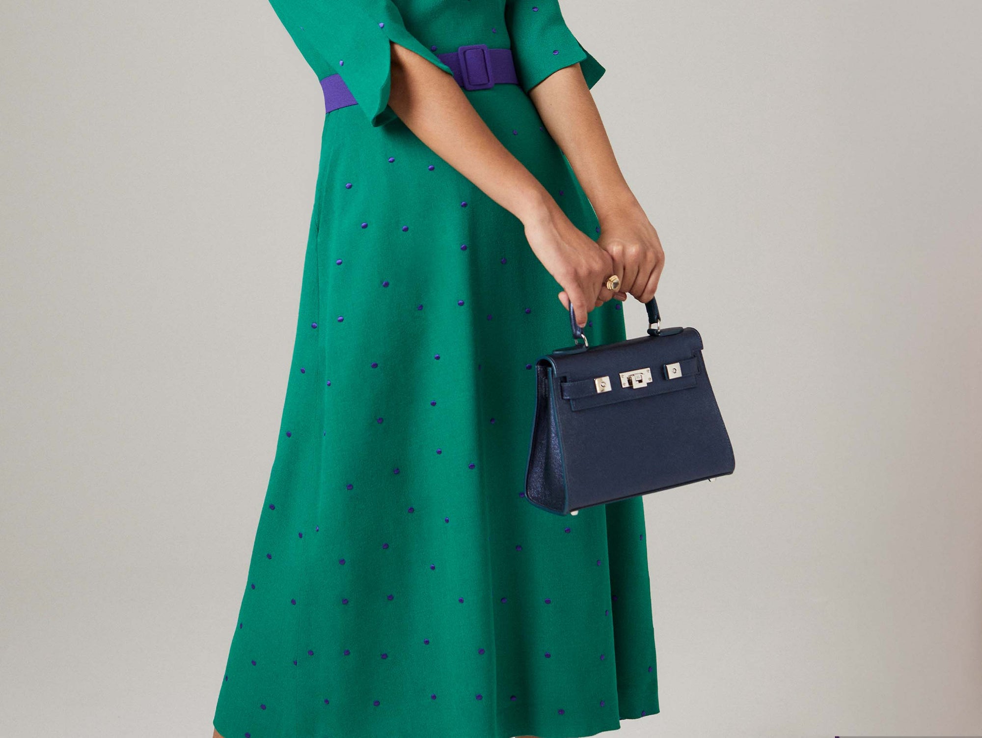 Person wearing a green dress with purple polka dots holding a Medium navy blue natural grain Italian leather handbag with a hint of silver sparkle  with silver hardware, a top handle and a detachable strap against a neutral background