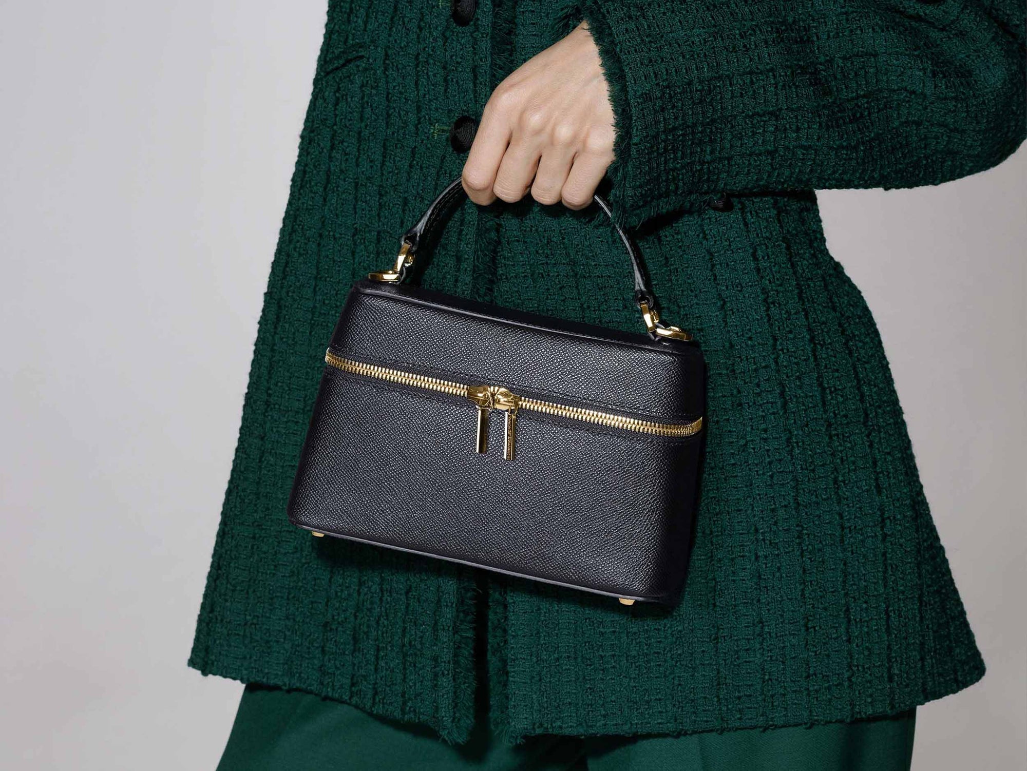 Person holding a black leather handbag with gold zipper by Lalage Beaumont against a plain background